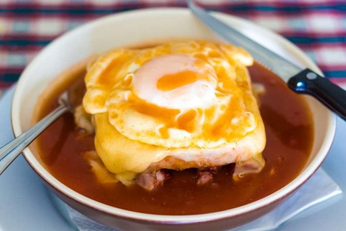 Un francesinha portugaise nappée de sauce tomate et surmontée d'un œuf au plat, un plat emblématique des habitudes alimentaires au Portugal.