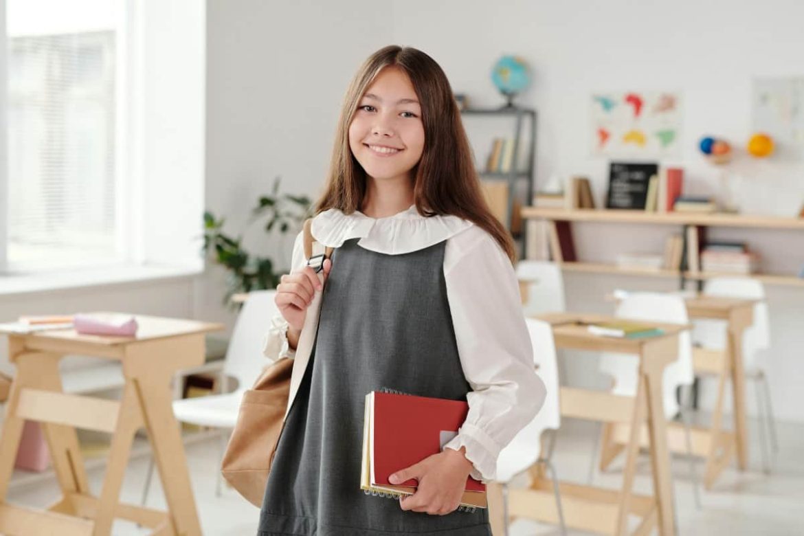 Une jeune étudiante portant un uniforme scolaire et un sac à dos tient ses livres en souriant dans une salle de classe, prête pour ses cours de piano à Nantes.