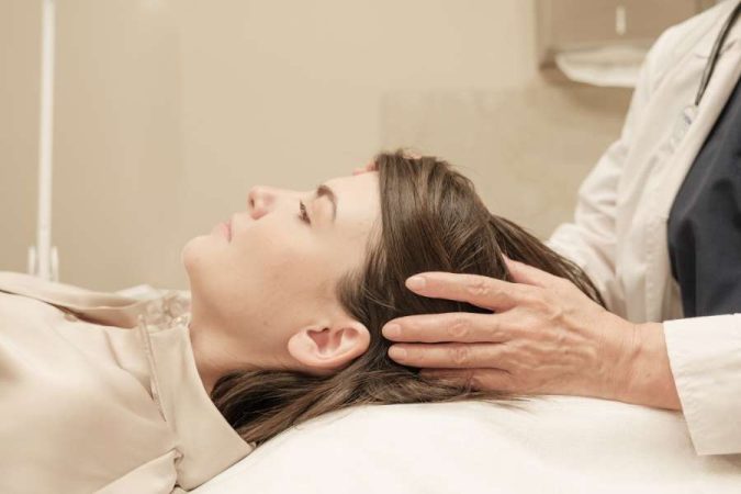 Une professionnelle examine et masse délicatement les cheveux d'une femme allongée lors d'un soin ciblé pour le gommage du cuir chevelu.